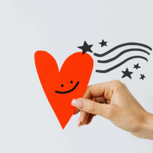 This image is decorative. It shows a hand holding a red cut out paper heart. On the heart is a hand drawn smily face. In the heart's wake there are stars and swoosh lines. This image represents how it can feel to give a recommendation or referral that works out.